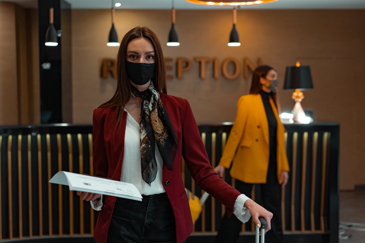 A woman in a mask stands at a hotel reception desk holding paper, welcoming service