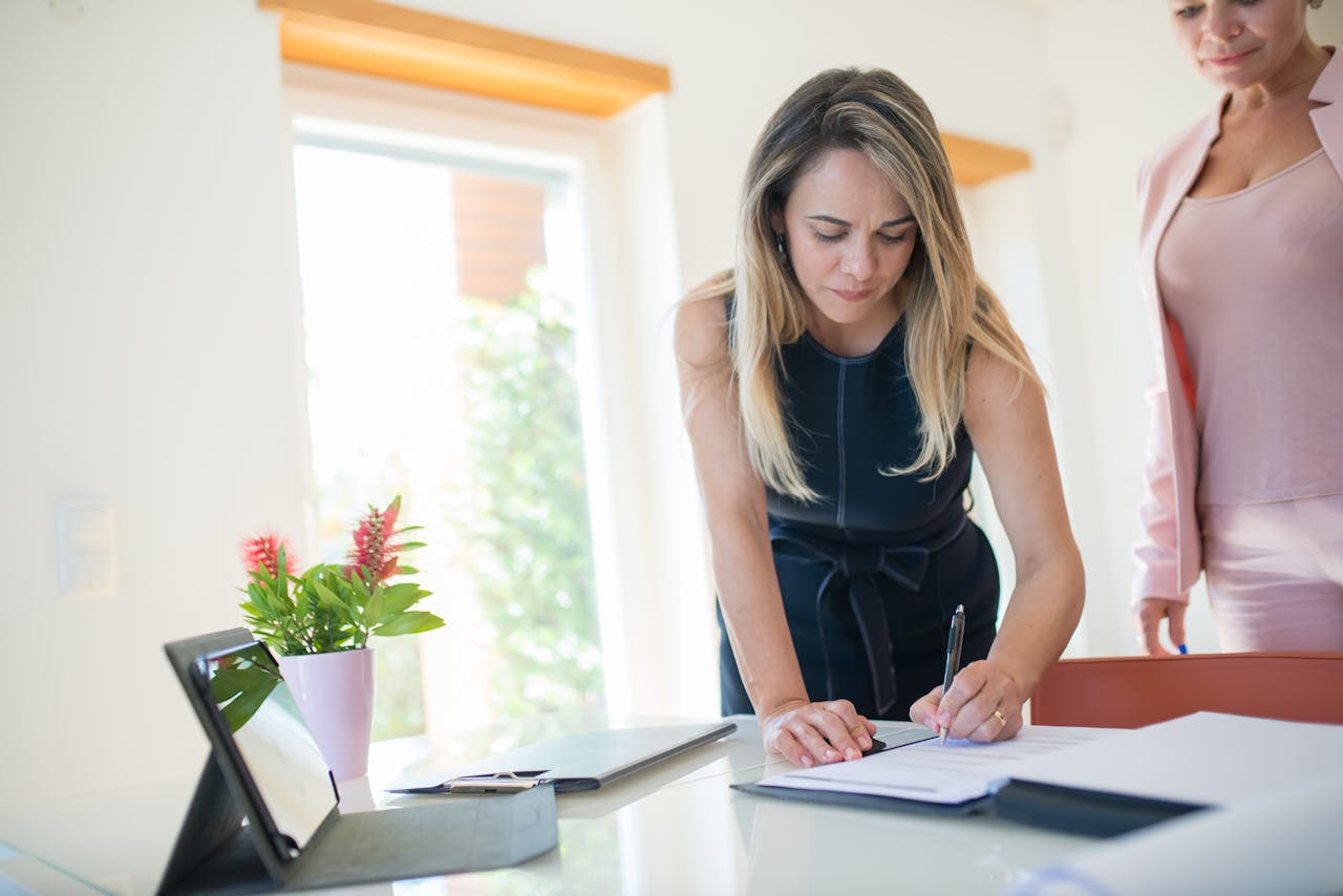 Businesswomen signing a contract at a bright office desk with documents and tablet.